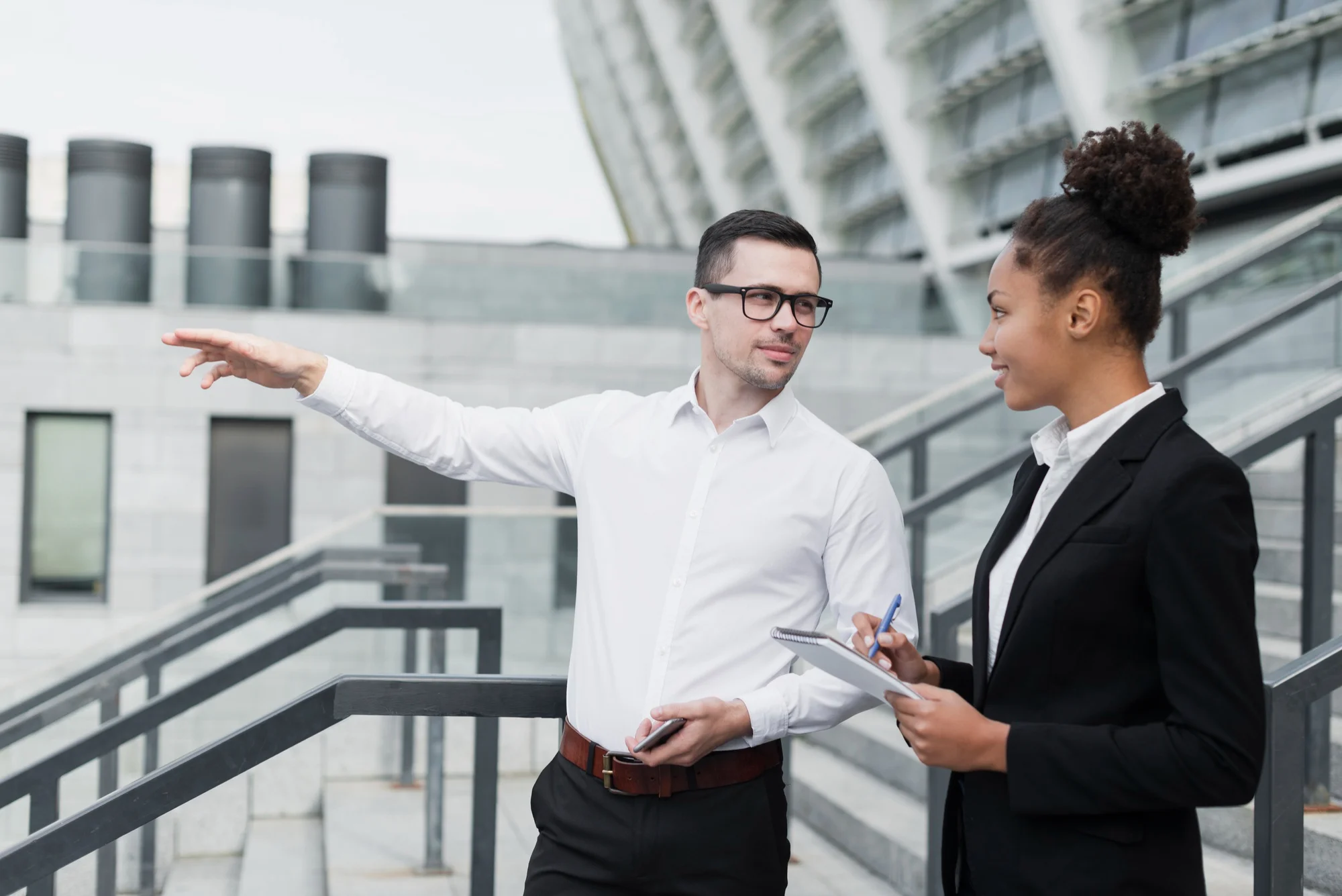 A businessman in a white shirt and glasses pointing toward a modern building while explaining growth insights to a female colleague taking notes.